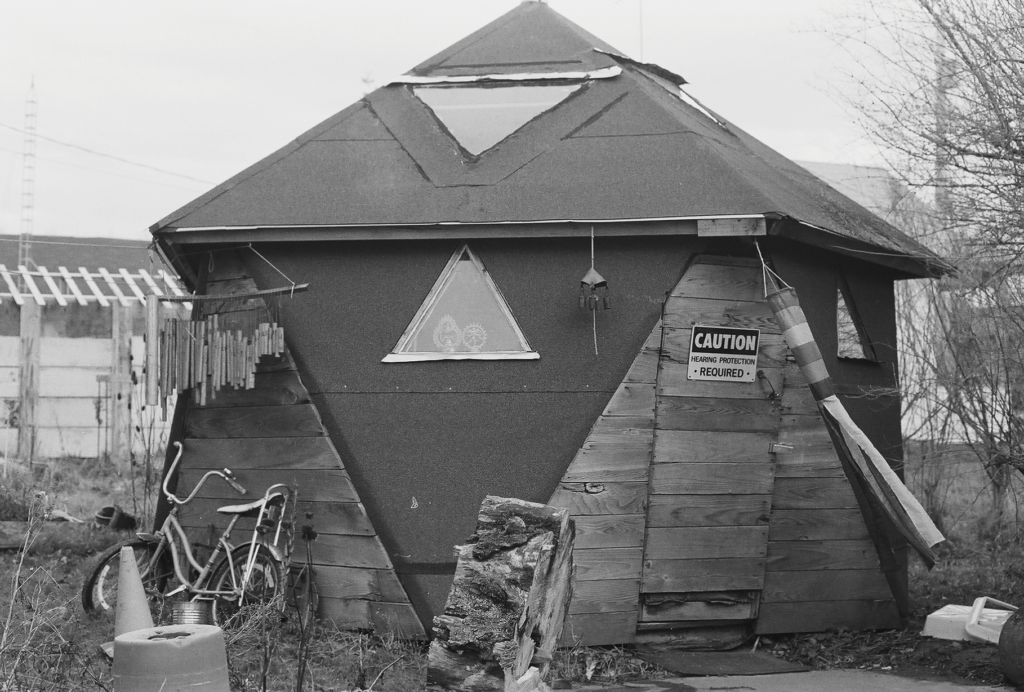 a large 1V geodesic dome. two windows and door are visible. one window has two hindu murtis in it. there is a large wind chime, and a child's bike leaned up against the side.